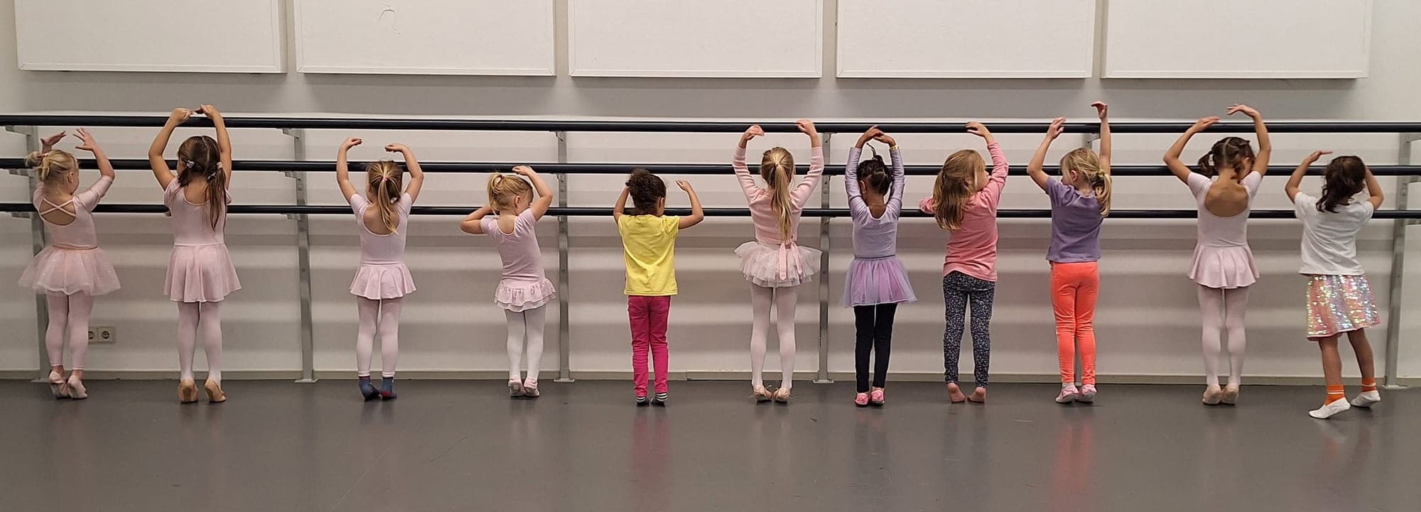 Een rij jonge danseresjes staat aan de barre in de balletzaal met de armen in een boog boven het hoofd
